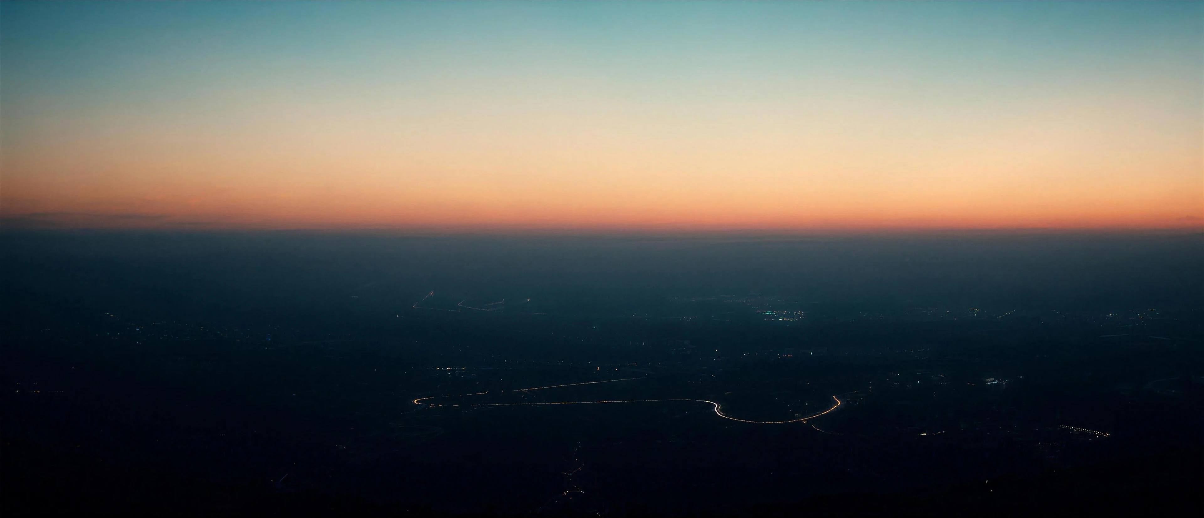 A valley seen from the air at dusk, one road drawn as a fine line of warm light winding between distant towns