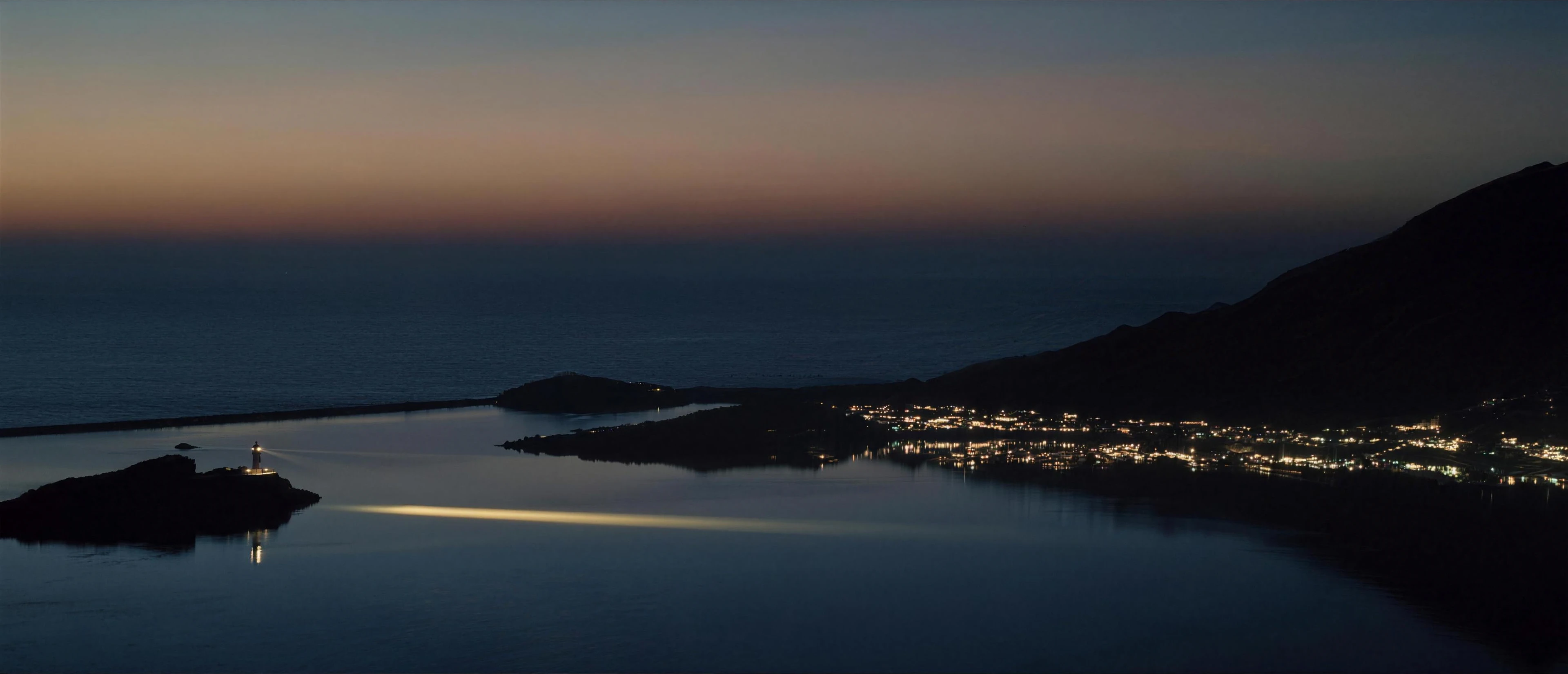 A lighthouse on a headland at dusk sends one long beam across still water toward the lit harbours of the far shore
