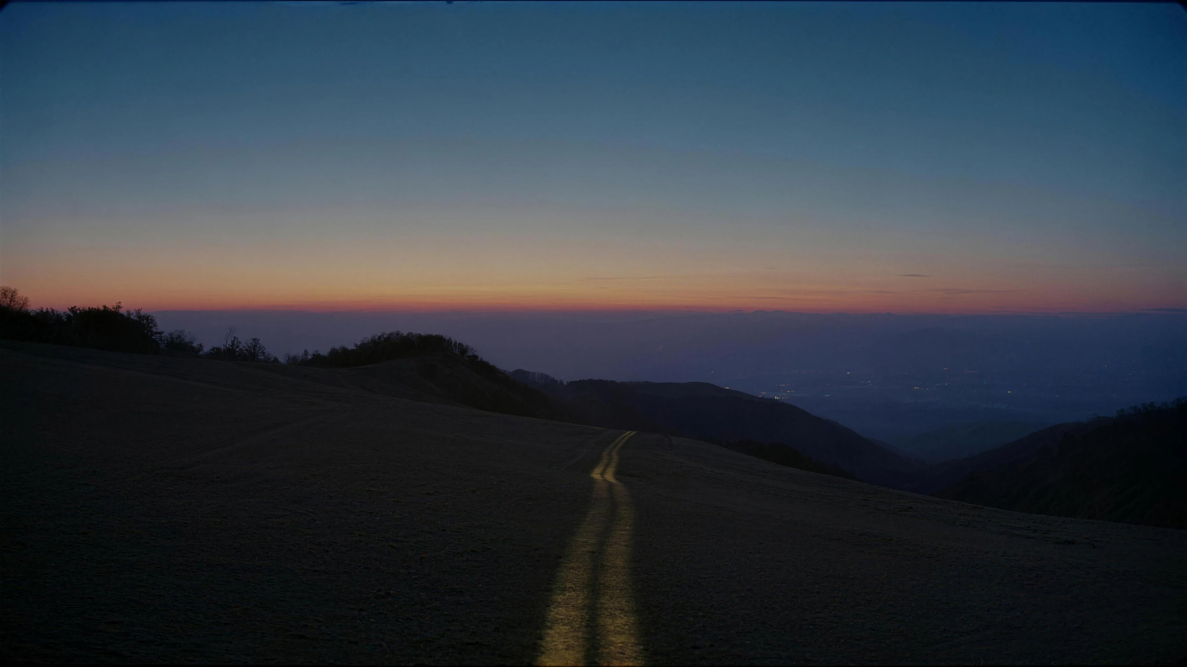 A grassy ridge at the last light of dusk, a lit path leading down into a valley of distant lights