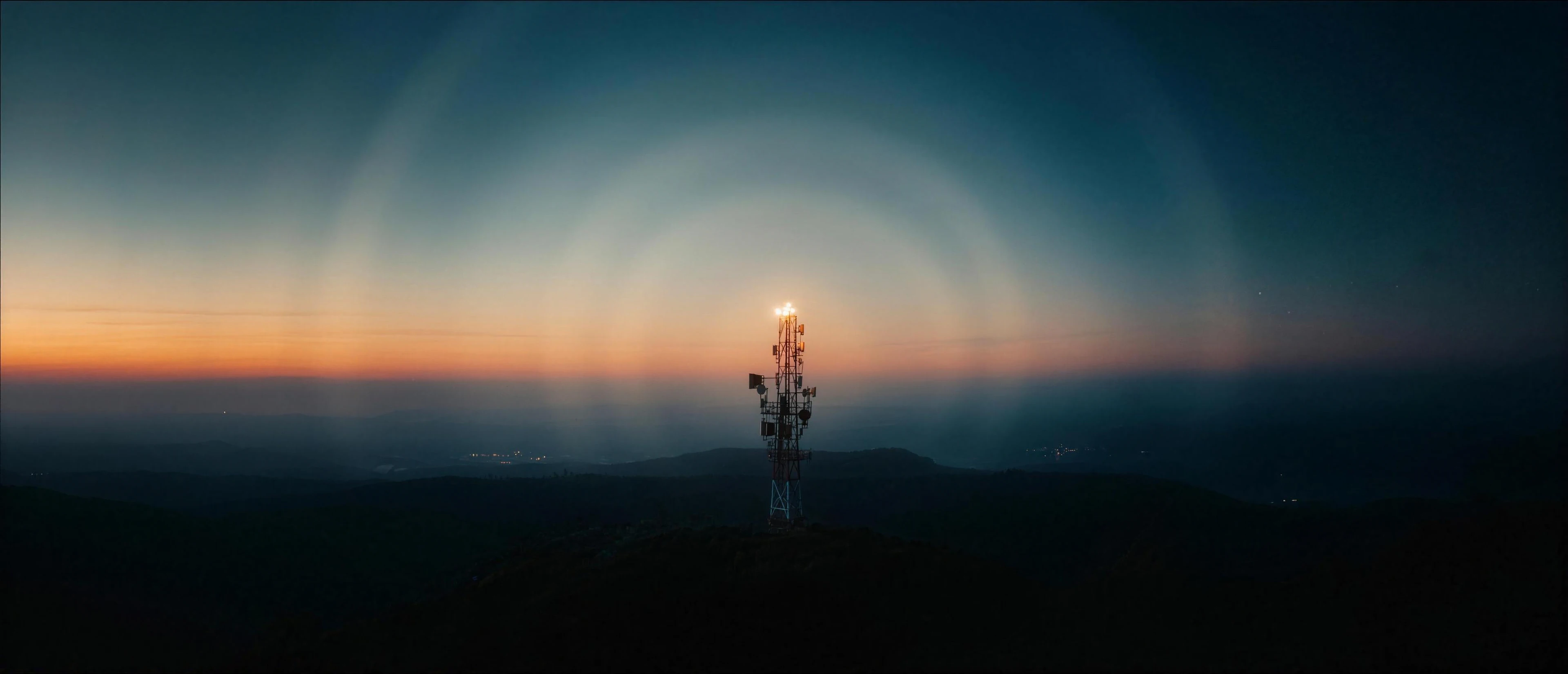 A broadcast tower on a dark hilltop at dusk, a warm beacon at its tip and rings of light spreading out over the hills